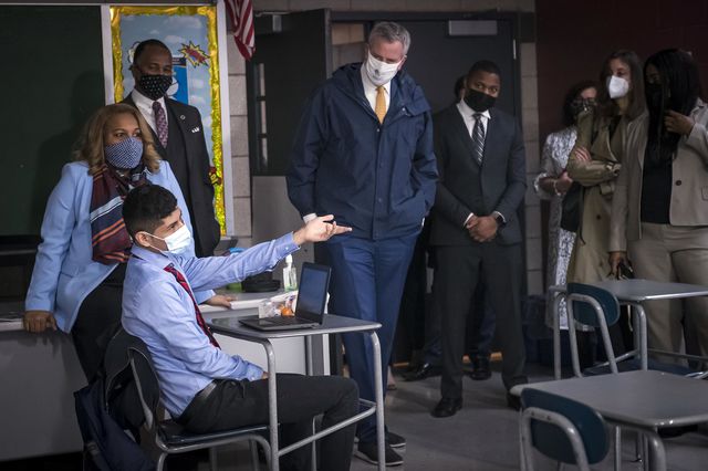 A student sitting in a desk while wearing a mask gesticulates while de BLasio, Porter, and others look on
